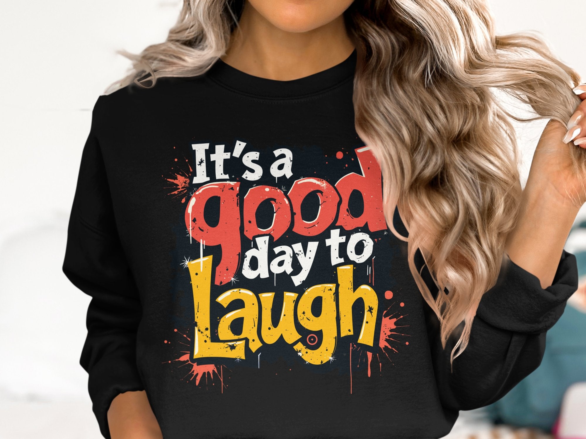 A person with long, wavy hair wears a Garment Graphics sweatshirt made from medium-heavy fabric. The black design features colorful text, Its a good day to laugh, with red, yellow, and white splatters, available in unisex sizes for everyone to enjoy.