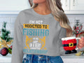 A person with long hair holds a Santa-themed mug, wearing a Garment Graphics gray sweatshirt that reads, Im not addicted to fishing. We are just in a very committed relationship, exuding its unisex style amid festive holiday decorations.