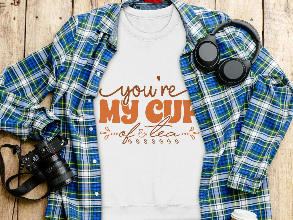 A white Youre My Cup of Tea t-shirt from Garment Graphics is displayed on a wooden surface, surrounded by a plaid shirt, headphones, camera, and coffee cup, making it perfect for tea enthusiasts and cozy sweatshirt lovers.