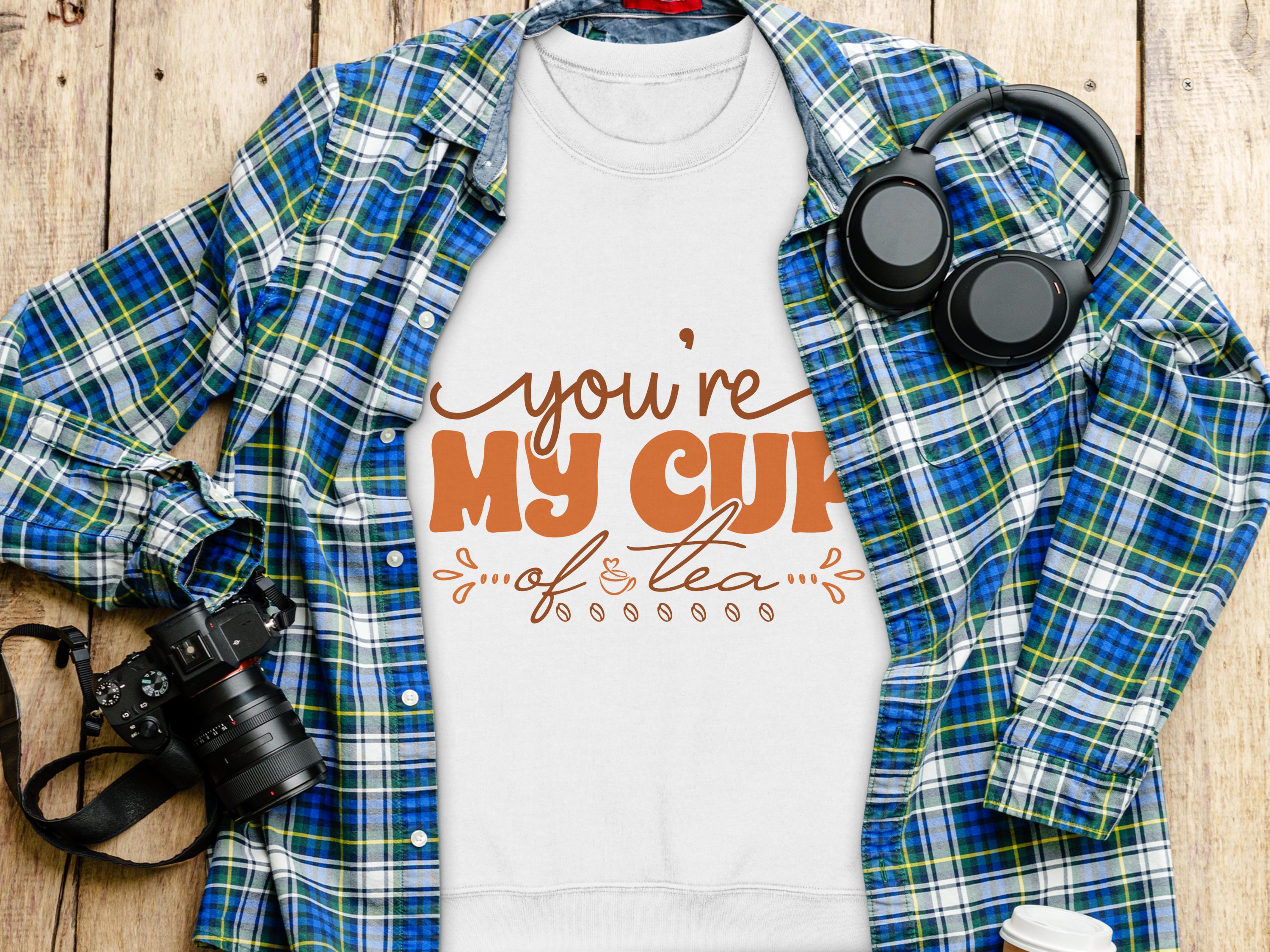 A white Youre My Cup of Tea t-shirt from Garment Graphics is displayed on a wooden surface, surrounded by a plaid shirt, headphones, camera, and coffee cup, making it perfect for tea enthusiasts and cozy sweatshirt lovers.