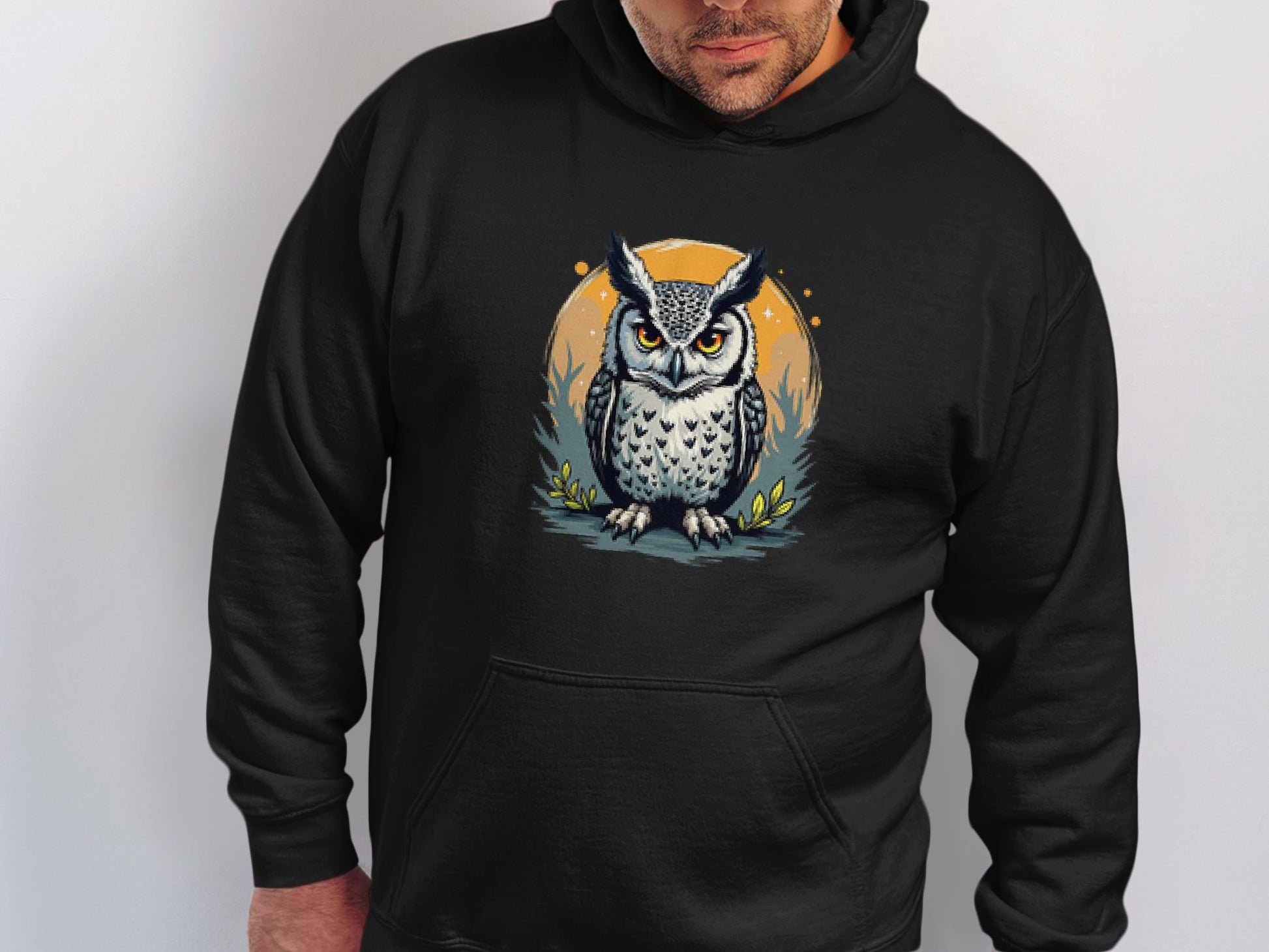 Someone is wearing a black hoodie from Garment Graphics Fierce Owl Illustration Hoodies, featuring a gray owl with large eyes grounded on an orange circle amid grass and small leaves. Its crafted from medium-heavy fabric for comfort.