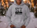 A person dons a gray Garment Graphics hoodie featuring Enjoy Classic Car and a vintage car illustration, standing in a softly lit snowy scene. This hoodie perfectly captures the community spirit of classic fit aficionados.