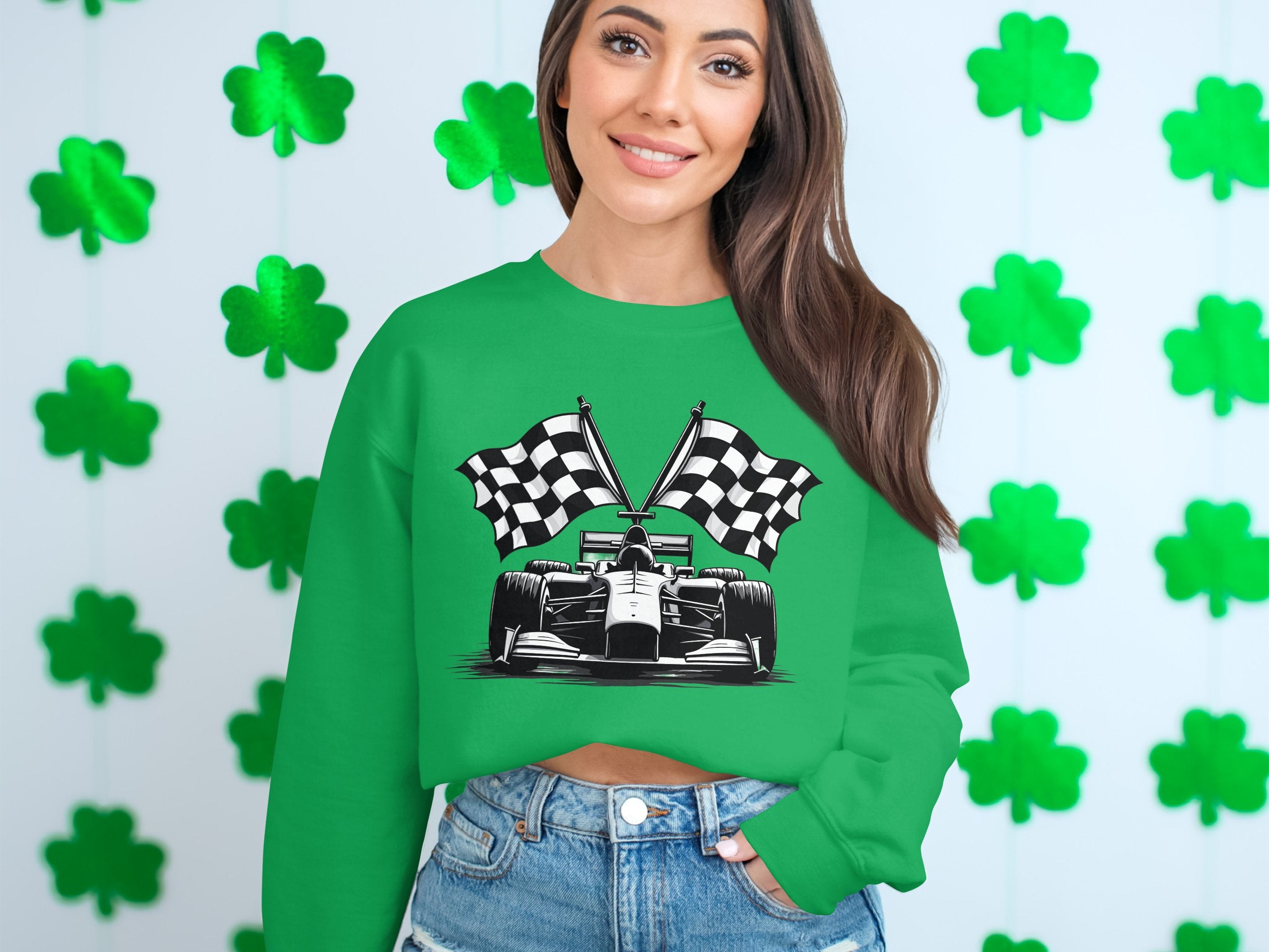 A woman stands smiling in front of a wall of green shamrocks, wearing Garment Graphics unique design—a green racing car sweatshirt with checkered flag print—and blue jeans. Her long hair cascades over one shoulder.