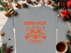 Garment Graphics displays a gray football sweatshirt with an orange Born to Play design on a wooden table, surrounded by Christmas decor—red and gold ornaments, pinecones, a star, and coffee. Perfect for young aspiring footballers, available in unisex sizing.