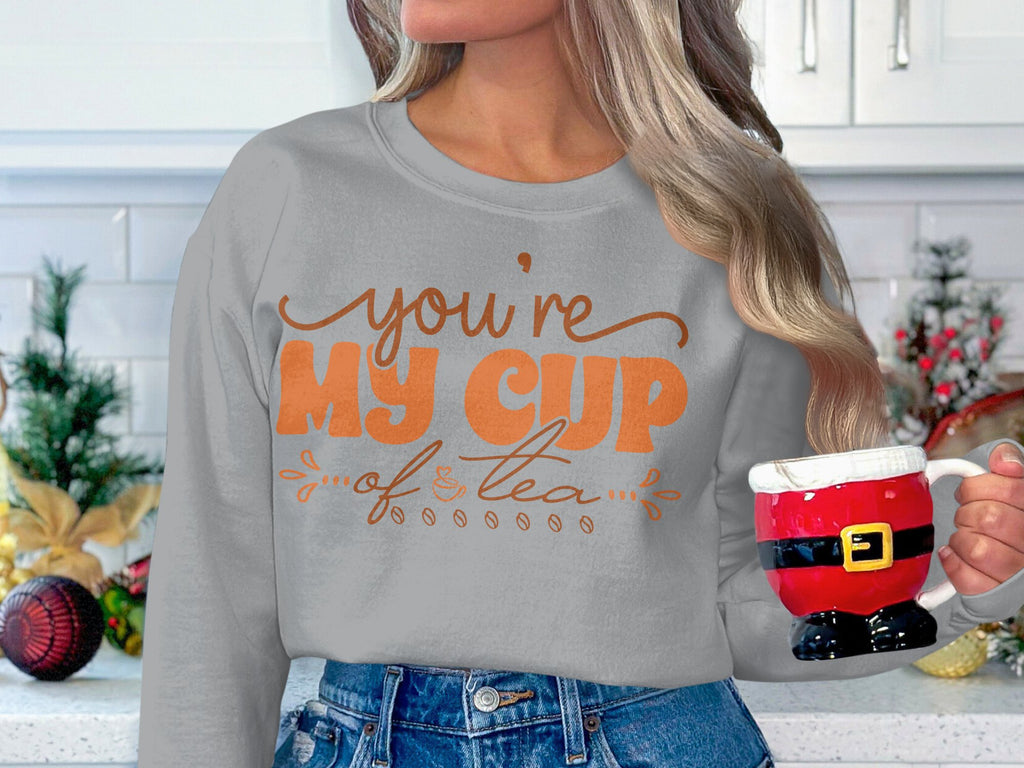 In a festive kitchen, someone holds a Santa-themed mug and wears Garment Graphics cozy sweatshirt with Youre My Cup of Tea in orange text. Holiday cheer fills the air with small trees and ornaments. The unisex sizing makes it an ideal festive favorite.