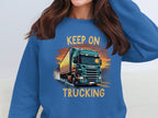 A Garment Graphics blue graphic sweatshirt features a green truck on a road, ideal for truck enthusiasts. The vibrant illustration has KEEP ON above and TRUCKING below. The persons face is hidden in the image.