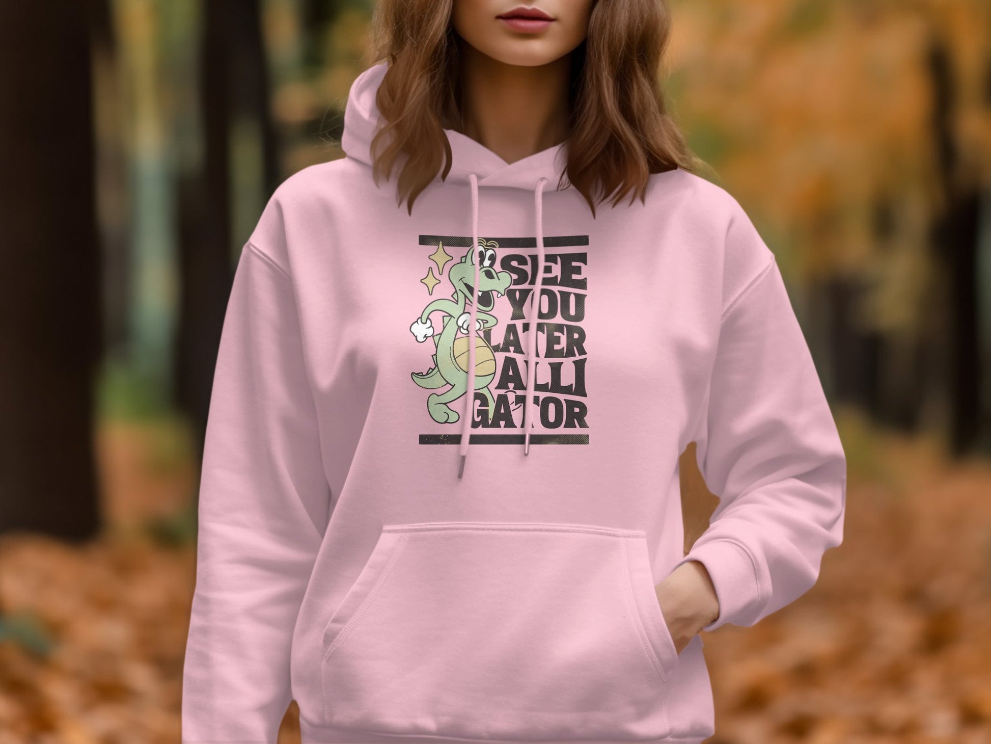 A person wears a classic fit Garment Graphics See You Later Alligator hoodie with a playful gator graphic. An out-of-focus forest backdrop features autumn-colored leaves, adding a seasonal vibe to the scene.