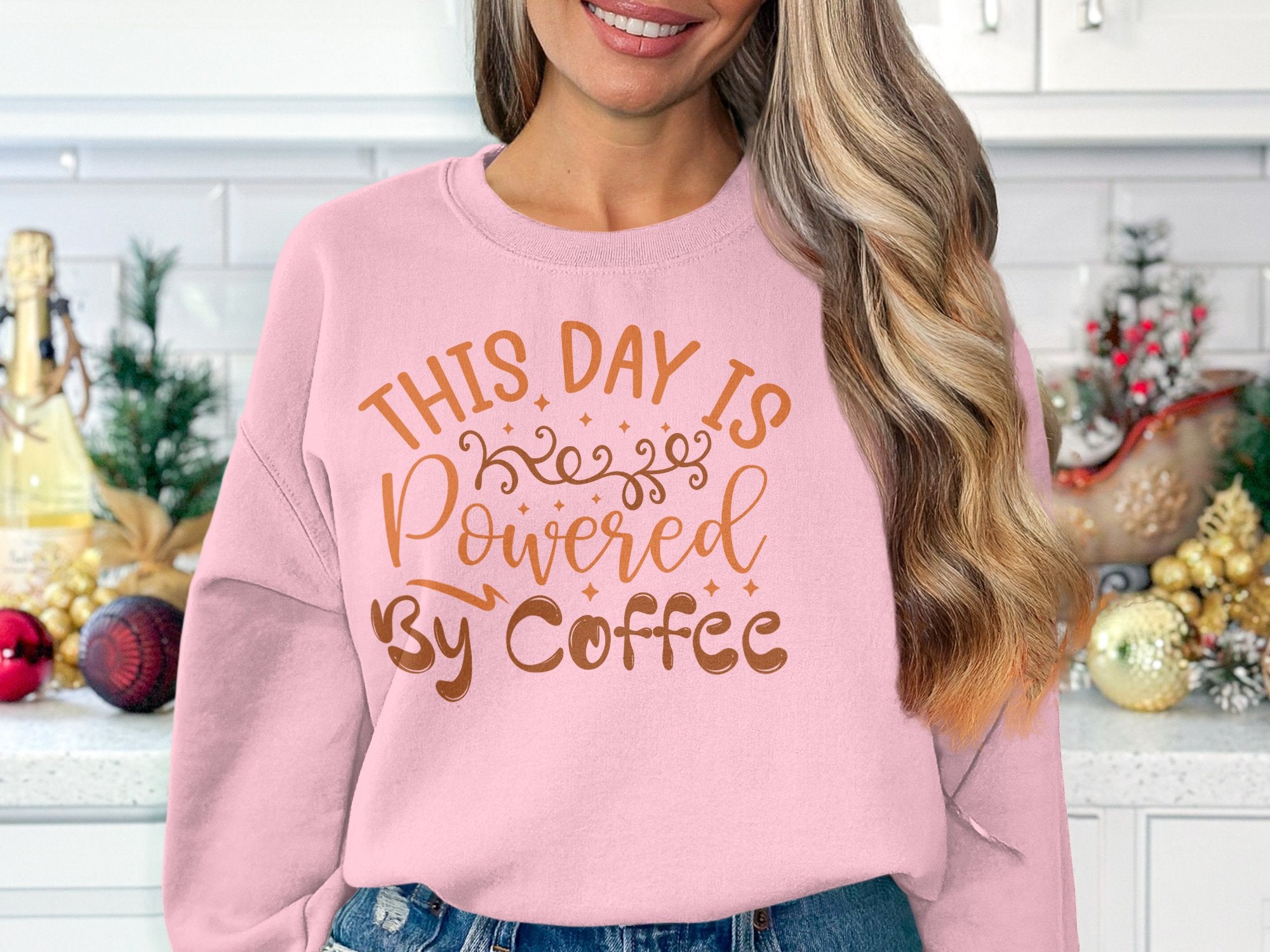 A woman wears a light pink sweatshirt by Garment Graphics with This Day is Powered by Coffee in brown lettering. She stands in front of a festively decorated kitchen counter adorned with holiday ornaments and greenery.