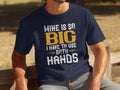 A person sits outdoors in a Garment Graphics fishing-themed navy tee with the humorous phrase, Mine is so big I have to use both hands, boldly printed in white and yellow. They relax on a wooden bench, enjoying the day.