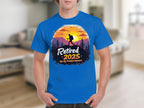 A person wears a blue sunset T-shirt by Garment Graphics featuring a hiker silhouette and the bold text, Retired 2025, Not My Problem Anymore, capturing the essence of retirement. The blurred room setting adds to the relaxed vibe.