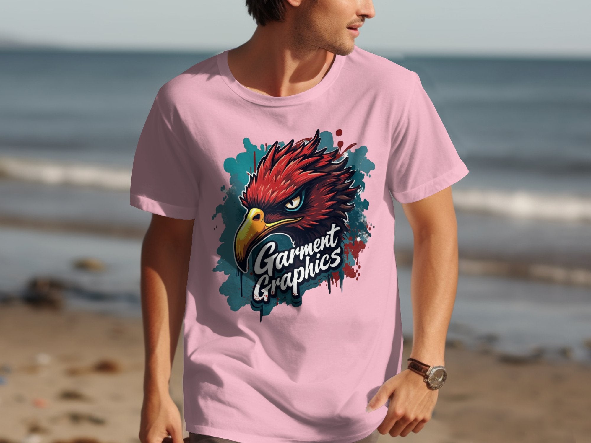A person in a pink Garment Graphics T-shirt with an eagle design strolls along the beach, framed by a stunning ocean backdrop.
