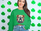 A woman wearing a Garment Graphics green sweatshirt with a bulldog design smiles, pairing it with denim shorts, as she stands against a white wall decorated with green shamrock garlands.