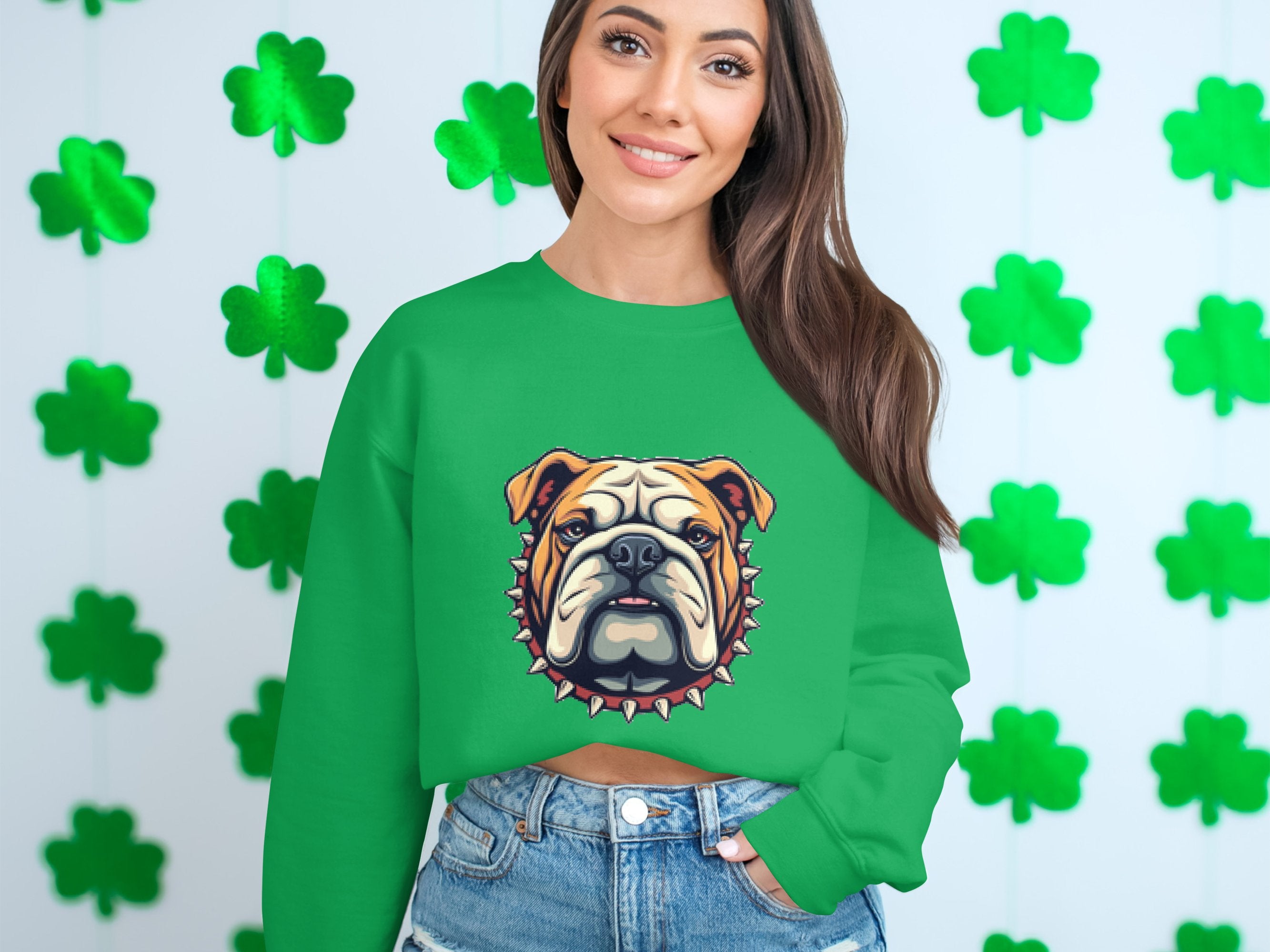 A woman wearing a Garment Graphics green sweatshirt with a bulldog design smiles, pairing it with denim shorts, as she stands against a white wall decorated with green shamrock garlands.