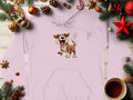 A Garment Graphics light pink cartoon dog hoodie rests on a wooden surface, ideal for dog lovers. Its surrounded by festive Christmas decor: pine branches, ornaments, pinecones, and a cozy cup of coffee.