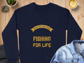 A Garment Graphics navy blue graphic sweatshirt with Father and Son Fishing for Life in bold yellow letters is showcased on a rustic wooden surface, paired with a steaming coffee cup, a vibrant plant, and a casual denim jacket.