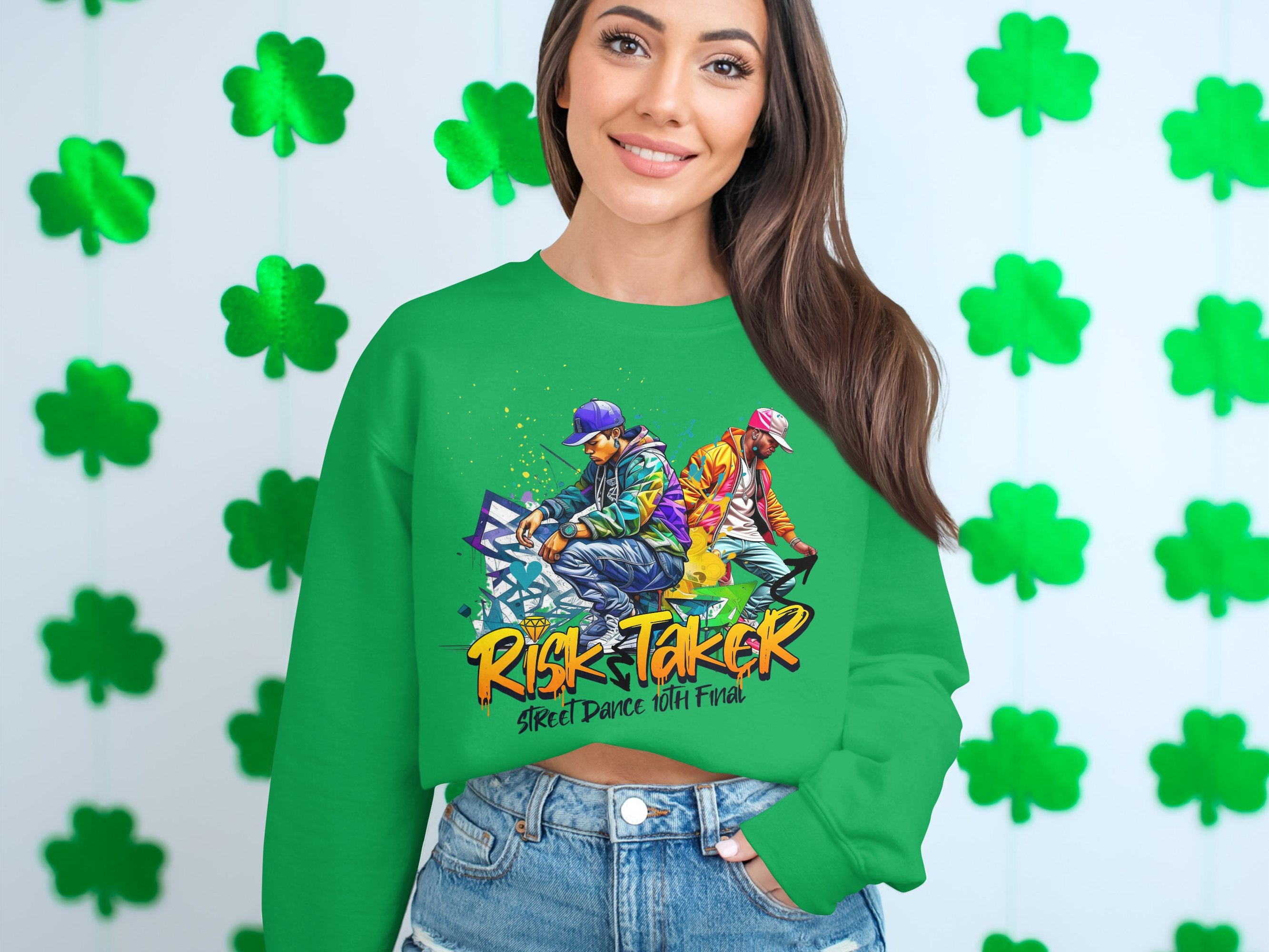 A woman confidently wears a Garment Graphics green sweatshirt featuring the Risk Taker Street Dance 10th Final logo and dynamic dancer graphics. Set against a backdrop of shamrocks on a white wall, she pairs it with blue jeans, capturing her street dance passion perfectly.