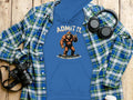 Garment Graphics presents a blue classic-fit hoodie with a Bigfoot graphic stating Admit It! Life Would Be Boring Without Me, surrounded by a plaid shirt, camera, headphones, and coffee cup on a wooden surface.