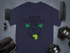 A navy Garment Graphics T-shirt saying Happy St. Patricks Day in black above three green shamrocks and two hearts sits on gray, giving a festive flair. Nearby are a smartwatch, smartphone, speaker, dumbbells, and black sneakers.