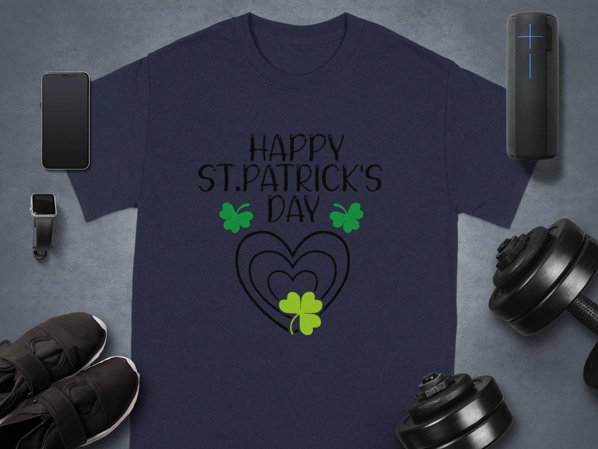 A navy Garment Graphics T-shirt saying Happy St. Patricks Day in black above three green shamrocks and two hearts sits on gray, giving a festive flair. Nearby are a smartwatch, smartphone, speaker, dumbbells, and black sneakers.