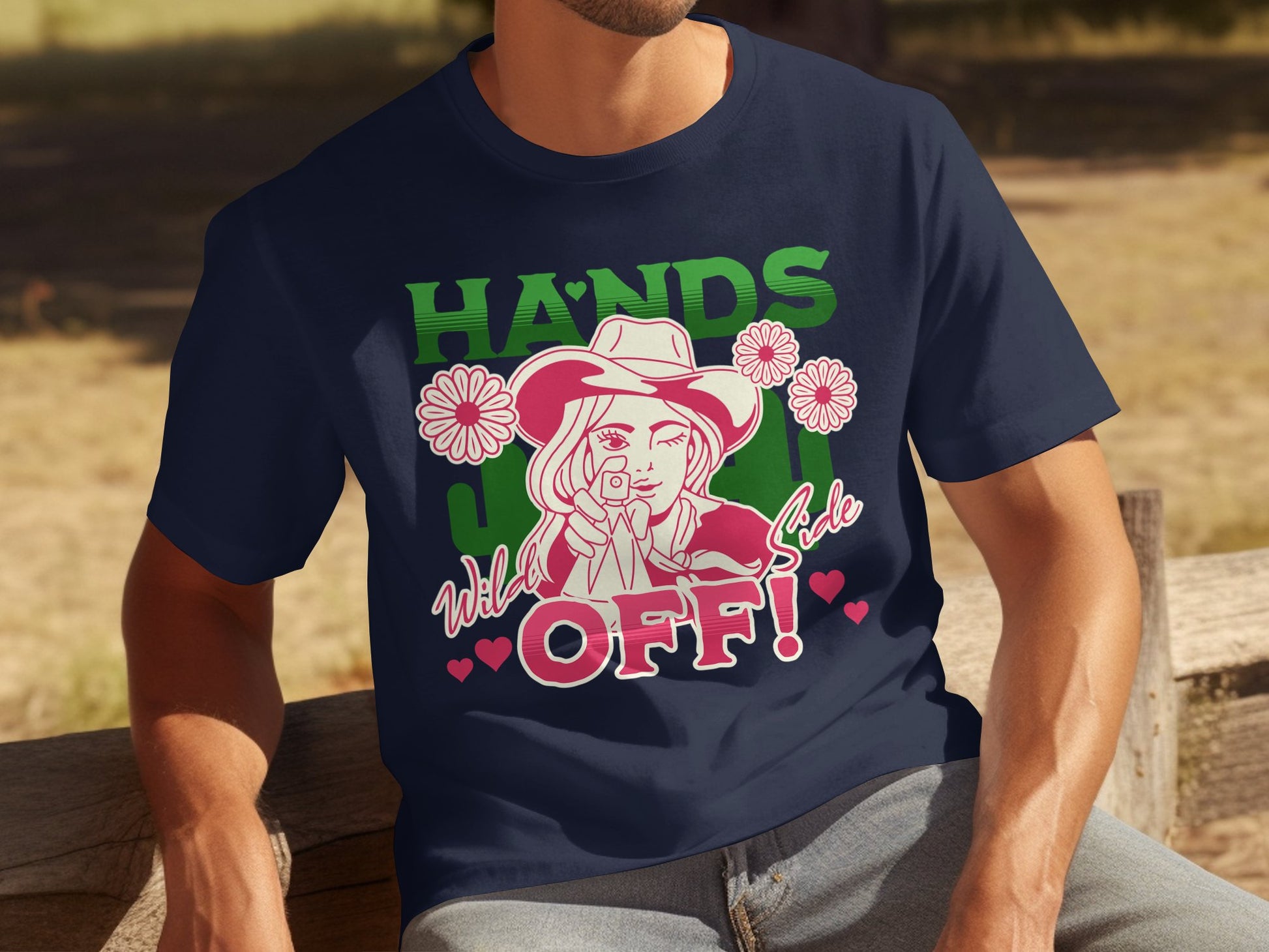 A man in a classic fit navy t-shirt, designed by Garment Graphics, features a bold cowboy hat design with flowers and the phrase HANDS WELL CODE OFF! in vibrant green, pink, and white. Hearts adorn the text as he sits on a wooden bench.
