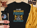 A black unisex sweatshirt by Garment Graphics features the text Im not addicted to fishing; we are just in a very committed relationship in diverse fonts and colors, surrounded by sunglasses, makeup, brushes, jewelry, a yellow blazer, and pink shoes on wood.