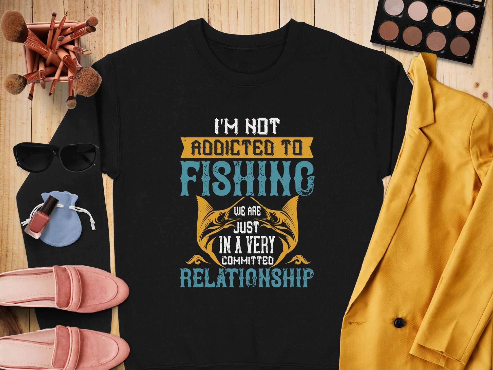 A black unisex sweatshirt by Garment Graphics features the text Im not addicted to fishing; we are just in a very committed relationship in diverse fonts and colors, surrounded by sunglasses, makeup, brushes, jewelry, a yellow blazer, and pink shoes on wood.