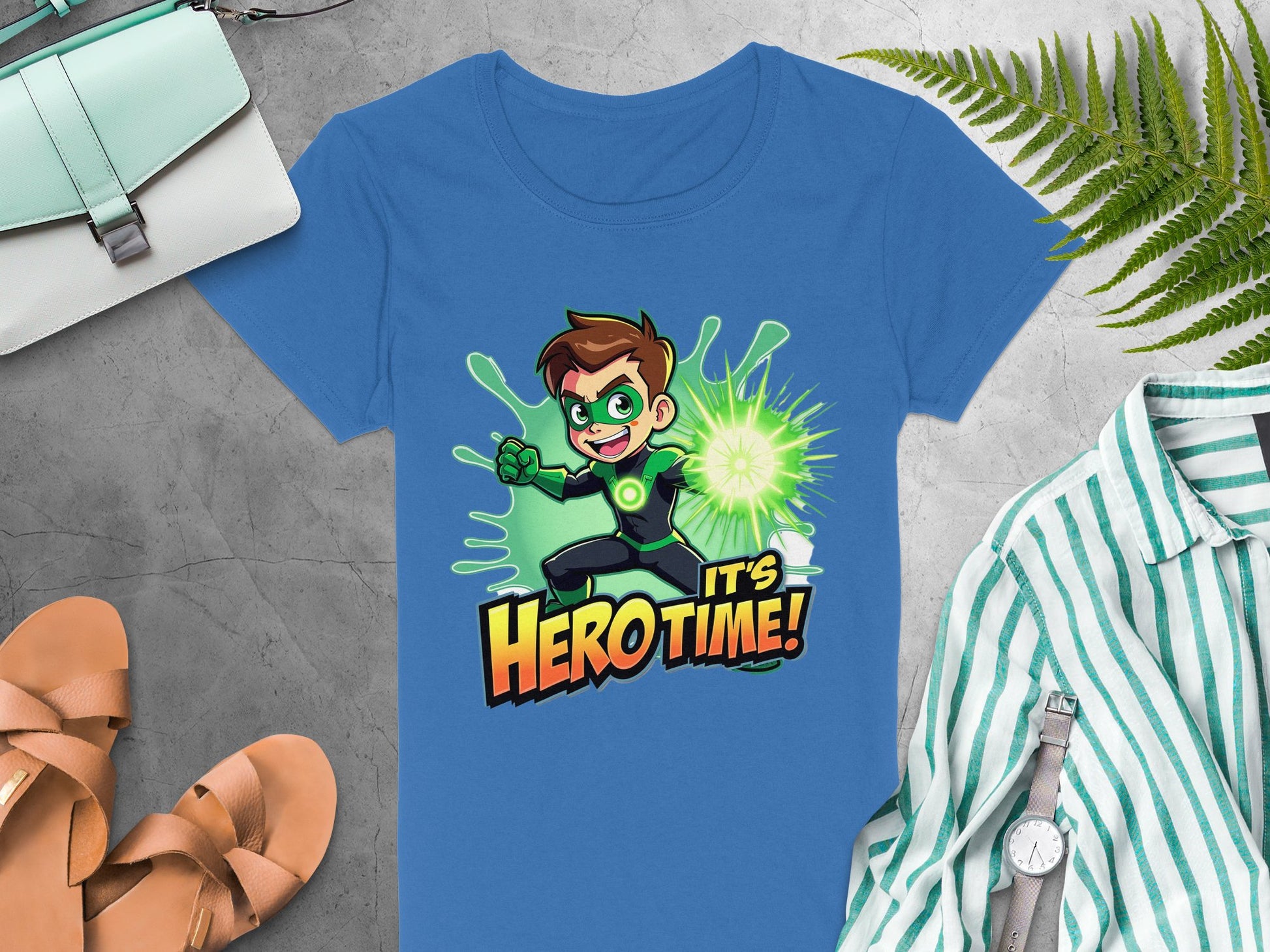 A blue Garment Graphics t-shirt with a green superhero, featuring an animated character and the text Its Hero Time!, is displayed among a light bag, brown sandals, and a green-striped shirt on a gray surface adorned with fern leaves.
