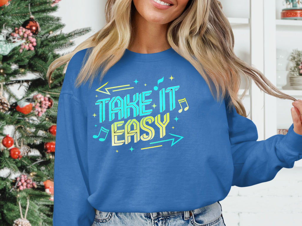 Someone is wearing a blue Garment Graphics sweatshirt with the Take It Easy graphic. The unisex, cozy attire pairs well with the festive indoor setting featuring a decorated Christmas tree. Only part of their face and hand, holding the sweatshirts hem, are visible.