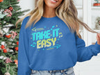 Someone is wearing a blue Garment Graphics sweatshirt with the Take It Easy graphic. The unisex, cozy attire pairs well with the festive indoor setting featuring a decorated Christmas tree. Only part of their face and hand, holding the sweatshirts hem, are visible.