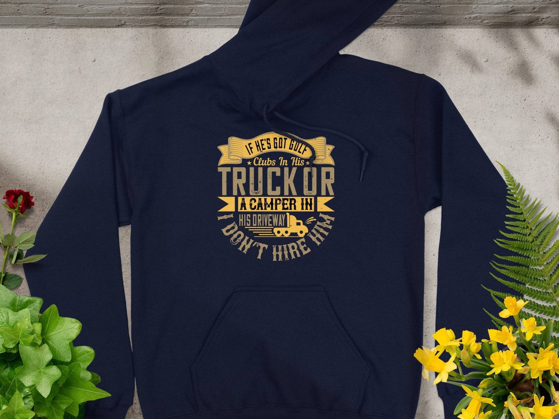 Garment Graphics navy golf hoodie with funny quote: If hes got cut-off clubs in his truck or a camper in his driveway, I dont hire him. An ideal gift displayed flat next to green and yellow leaves on light gray.