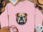 A Garment Graphics pink sweatshirt with a bulldog and spiked collar design is on a wooden surface, surrounded by a notebook, sandals, sun hat with sunglasses, camera, and brown bag strap. This stylish unisex piece suits everyone.