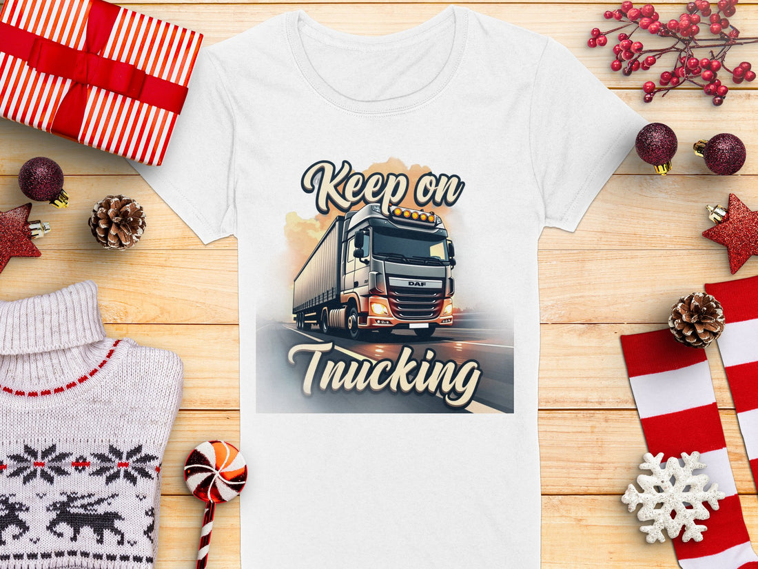 Garment Graphics white T-shirt with a truck graphic and Keep On Trucking text is displayed on wood, surrounded by striped gift boxes, pine cones, a snowflake, candy cane, and sweater.