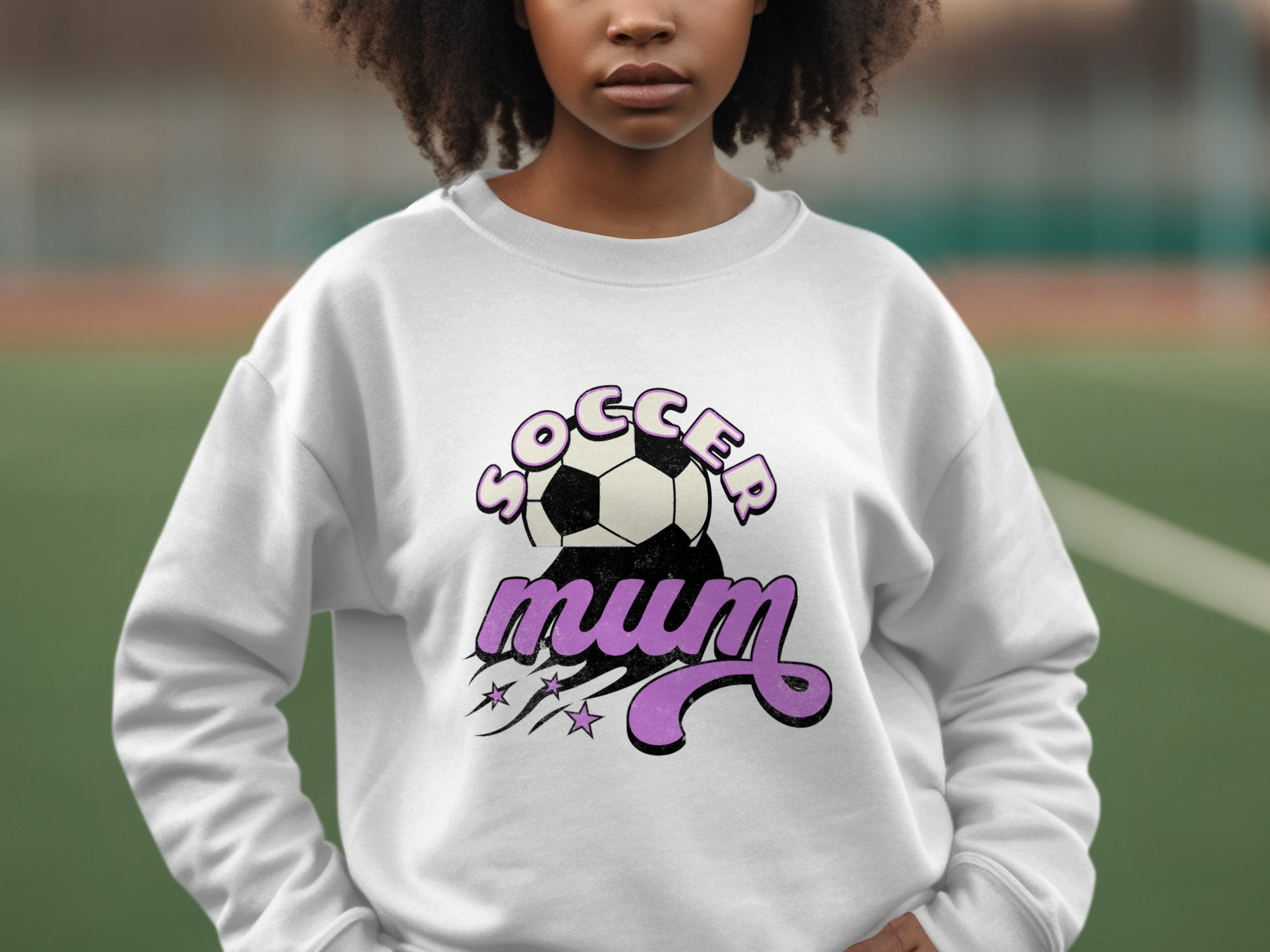 A person on a sports field wears a white Garment Graphics sweatshirt with a vintage Soccer Mum design, showcasing a soccer ball and purple stars.