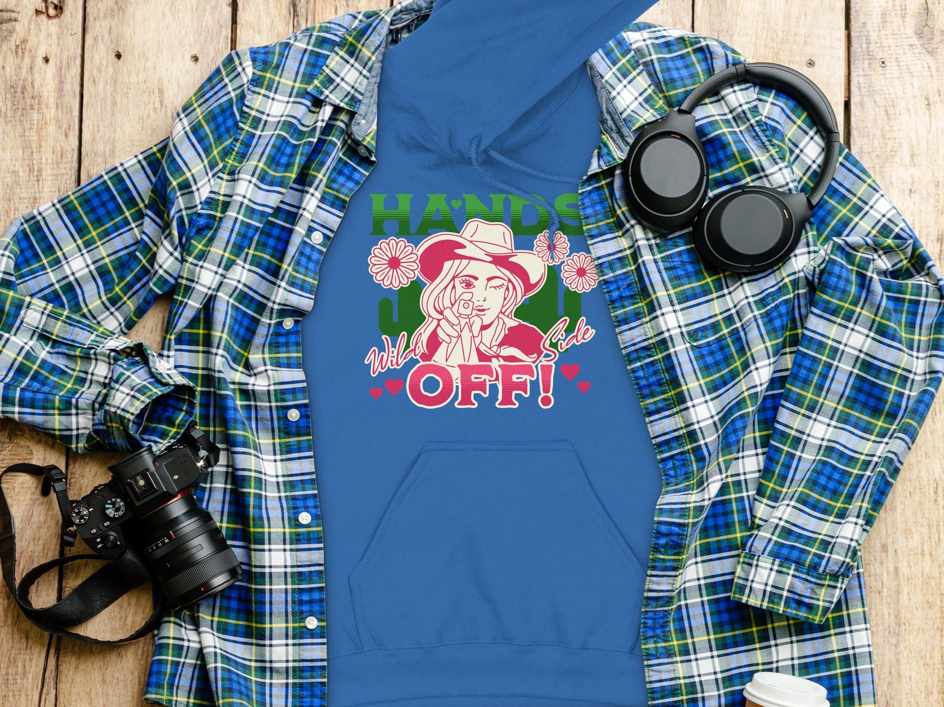 A blue Garment Graphics Bold Statement Hoodie with a graphic of a person in a hat, flowers, and Hands Off! text in vivid red and green. Styled on wood with a plaid shirt, black headphones, and camera for an adventurous look.