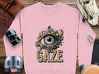 Garment Graphics offers a pink graphic sweatshirt with an intricate eye design and GAZE beneath it. This unisex piece lies on a wooden surface, styled with a camera, gloves, and binoculars for an adventurous feel.