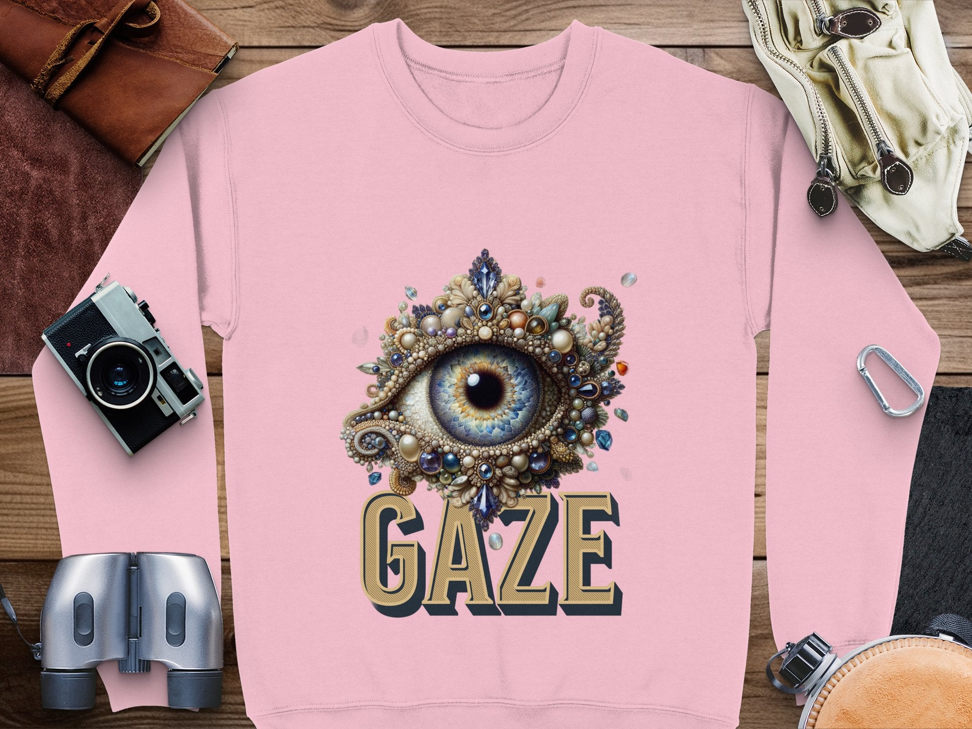 Garment Graphics offers a pink graphic sweatshirt with an intricate eye design and GAZE beneath it. This unisex piece lies on a wooden surface, styled with a camera, gloves, and binoculars for an adventurous feel.