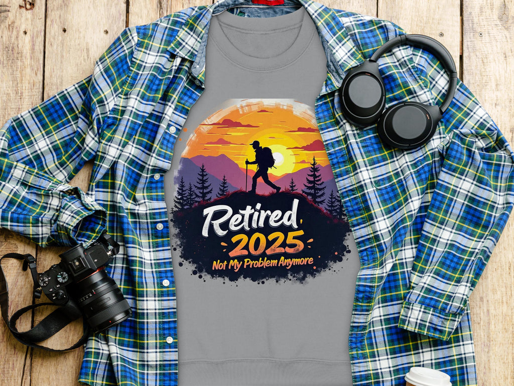 Garment Graphics unisex gray T-shirt features a hiker design with a sunset and trees, displaying Retired 2025 Not My Problem Anymore. Its styled over a checkered shirt, next to headphones, a camera, and a coffee cup on wood.