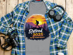 Garment Graphics unisex gray T-shirt features a hiker design with a sunset and trees, displaying Retired 2025 Not My Problem Anymore. Its styled over a checkered shirt, next to headphones, a camera, and a coffee cup on wood.