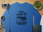 A Garment Graphics blue sweatshirt featuring a vintage car print, capturing classic automotive style, rests on wood. Nearby are a camera, lens, chopped cucumber bowl, classic London City map, book, and burlap bag.