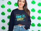 A person with long hair stands before a wall with green shamrocks, wearing a unisex sweatshirt by Garment Graphics, featuring the Take It Easy graphic in colorful letters. With one hand in their pocket, they smile casually at the camera.