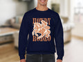 Wearing a Garment Graphics Howdy Rodeo Sweatshirt in navy blue, this person radiates cowboy spirit with a cowboy-on-horse graphic. The bold Howdy Rodeo text pops against the indistinct indoor background, an ideal choice for rodeo lovers.