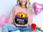 A person relaxes with red rose and pink heart-shaped balloons, wearing a Garment Graphics retirement sweatshirt. The unisex sizing design reads Retired 2025 - Not My Problem Anymore, showcasing a hiker silhouette against a sunset and trees.