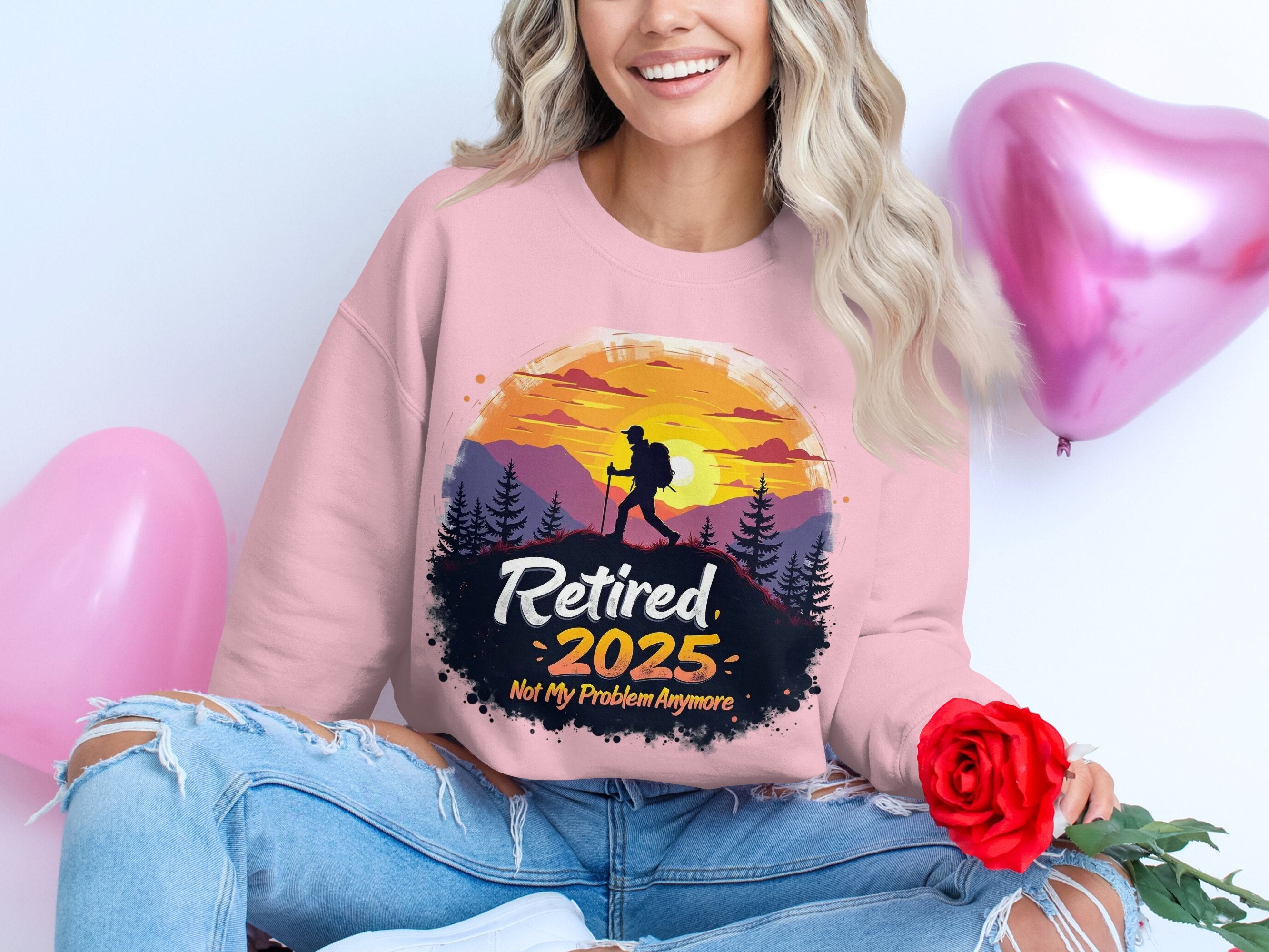 A person relaxes with red rose and pink heart-shaped balloons, wearing a Garment Graphics retirement sweatshirt. The unisex sizing design reads Retired 2025 - Not My Problem Anymore, showcasing a hiker silhouette against a sunset and trees.