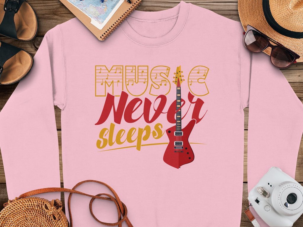 A trendy Garment Graphics pink sweatshirt sits on a wooden surface, ideal for music lovers. It features the Music Never Sleeps Guitar with yellow and red text and a striking red electric guitar. Nearby are a hat, sunglasses, sandals, notepad, camera, and purse.