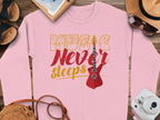 A trendy Garment Graphics pink sweatshirt sits on a wooden surface, ideal for music lovers. It features the Music Never Sleeps Guitar with yellow and red text and a striking red electric guitar. Nearby are a hat, sunglasses, sandals, notepad, camera, and purse.