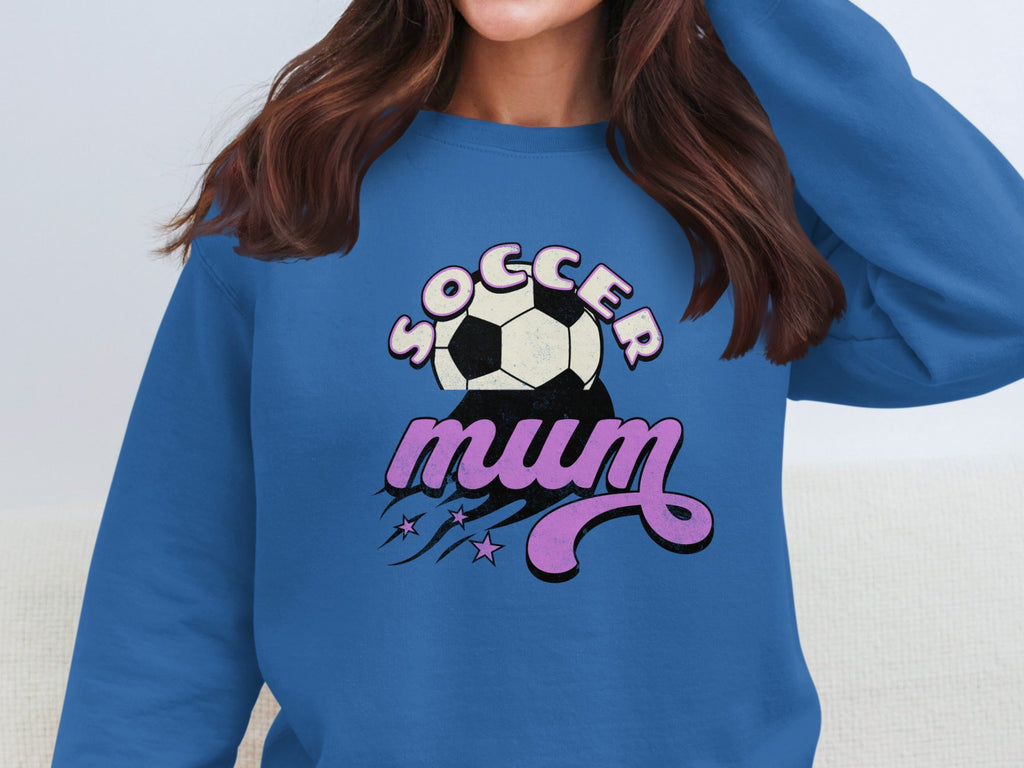 A woman wears a Garment Graphics vintage-style blue Soccer Mum sweatshirt adorned with pink and white text, soccer ball, and star accents. Her face is hidden to highlight the design.