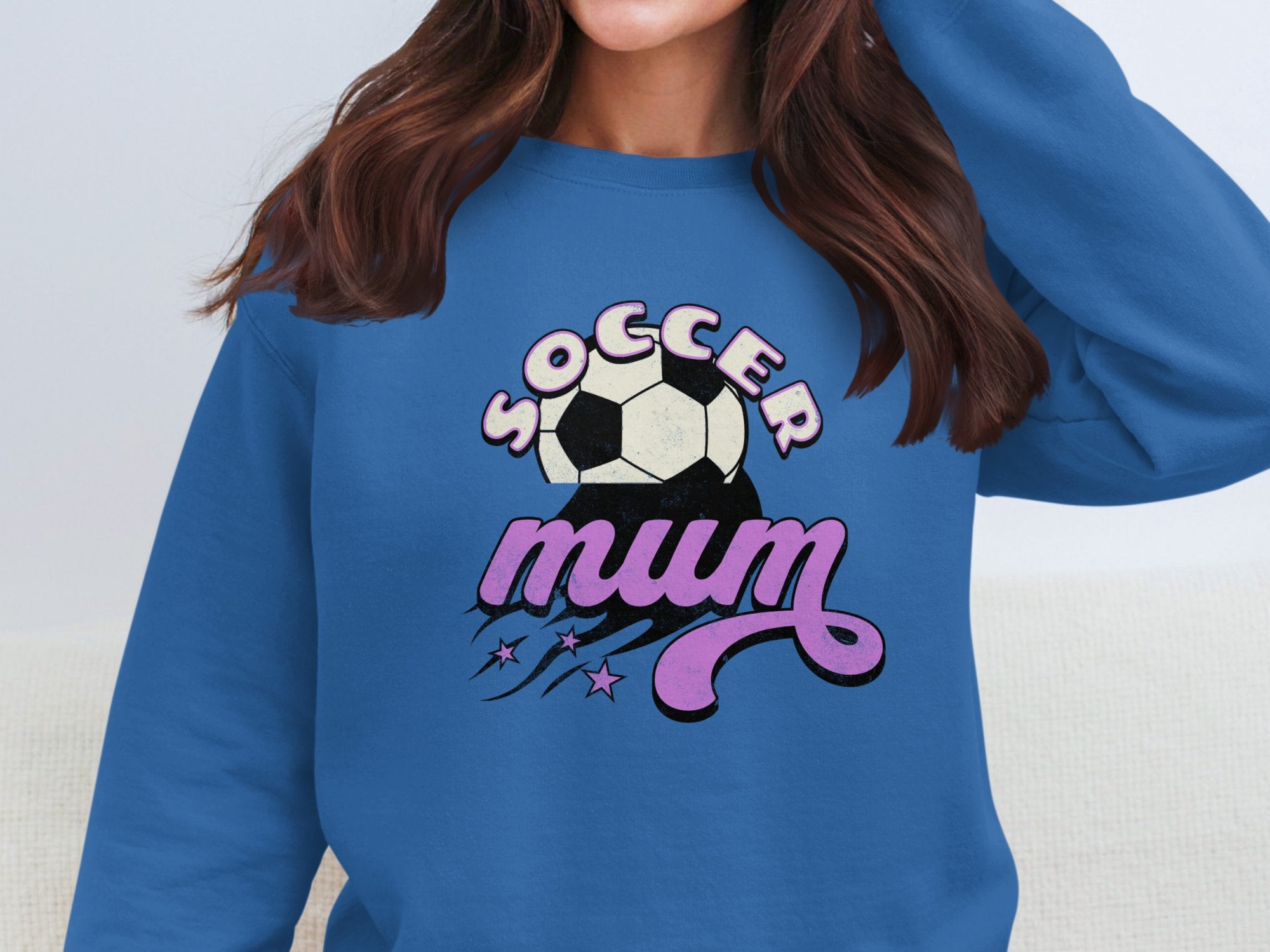 A woman wears a Garment Graphics vintage-style blue Soccer Mum sweatshirt adorned with pink and white text, soccer ball, and star accents. Her face is hidden to highlight the design.