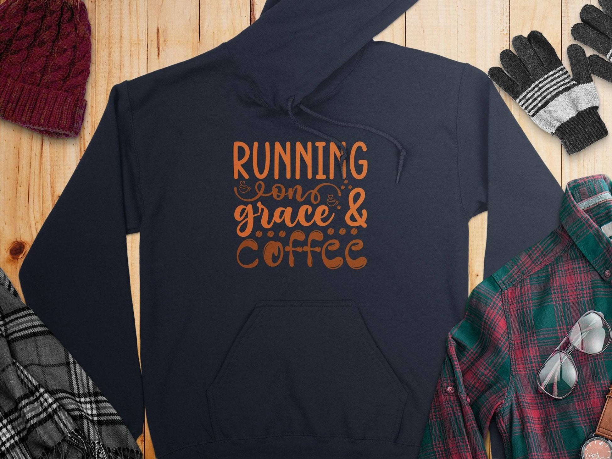 A navy classic fit hoodie from Garment Graphics with Running on Grace & Coffee printed in orange pairs perfectly with a burgundy knit hat, striped gloves, plaid scarf, and green and red plaid shirt on a wooden surface.