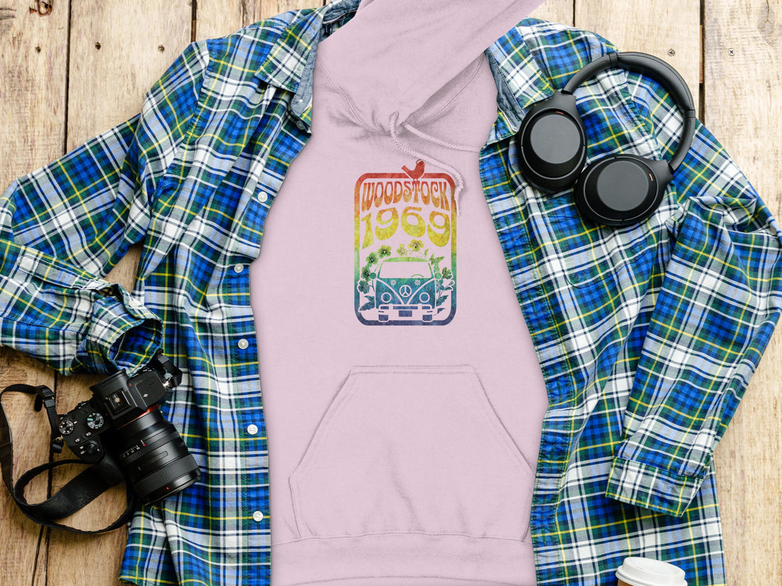 A person dons a retro black hoodie by Garment Graphics featuring a vintage Woodstock 1969 print. The design has a gradient with a car, flowers, and bird, against a plain white backdrop to emphasize its classic style.