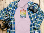 The Garment Graphics retro pink hoodie featuring a vintage Woodstock 1969 design is displayed on wood, accompanied by a blue-green plaid shirt, headphones, a camera, and a take-out coffee cup, encapsulating the essence of timeless graphic print hoodies.